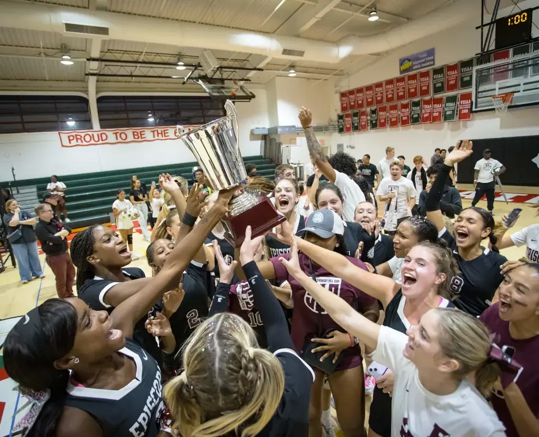 A girls' sports team celebrates a championship victory in a gymnasium, lifting a large silver trophy together. 