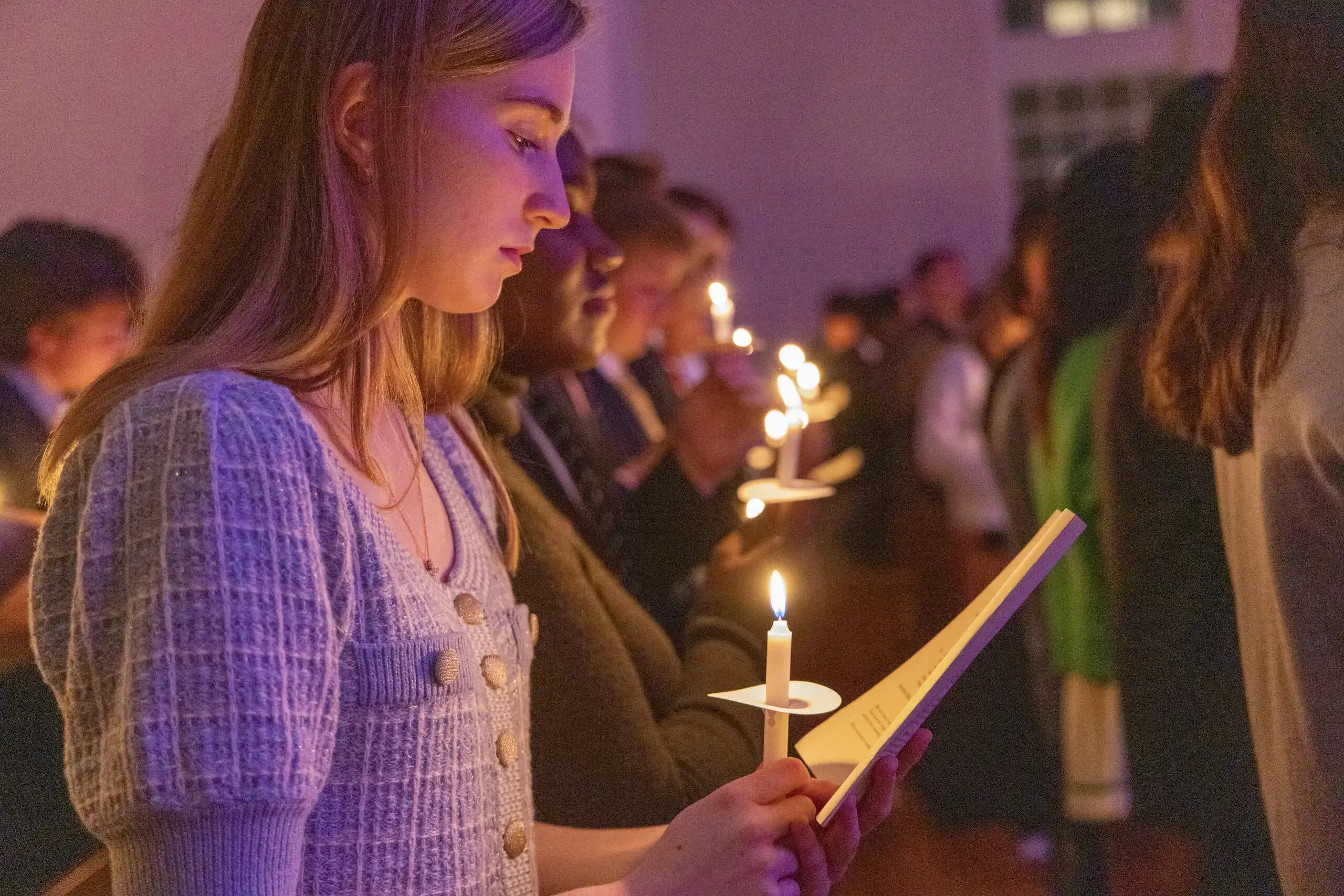 A young woman holds a lit candle and a program during a candlelight ceremony. She is surrounded by other participants holding candles in a dimly lit space, creating a warm, reflective atmosphere.