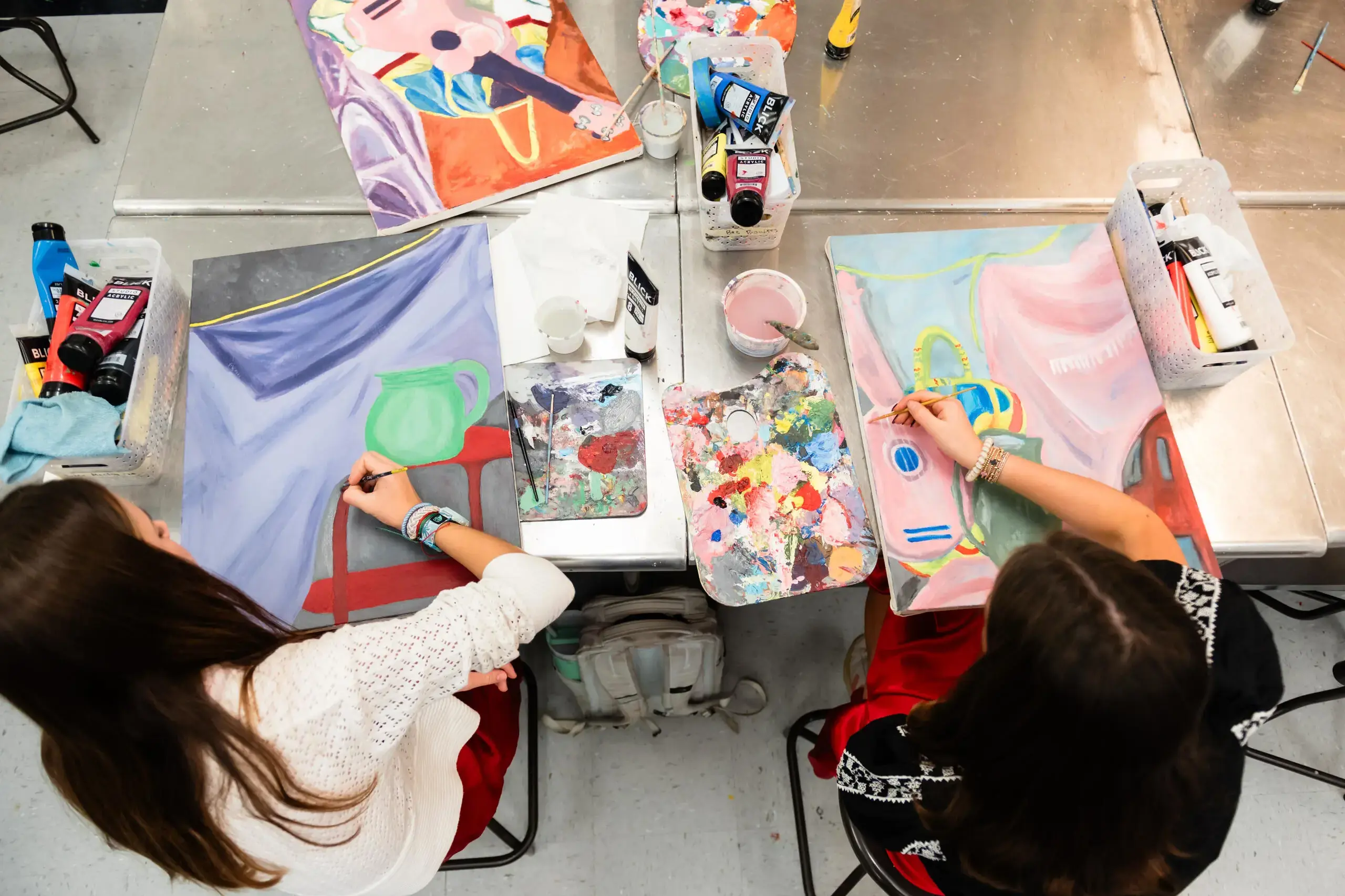 Two students sit at a table in an art classroom, each working on a colorful painting. 