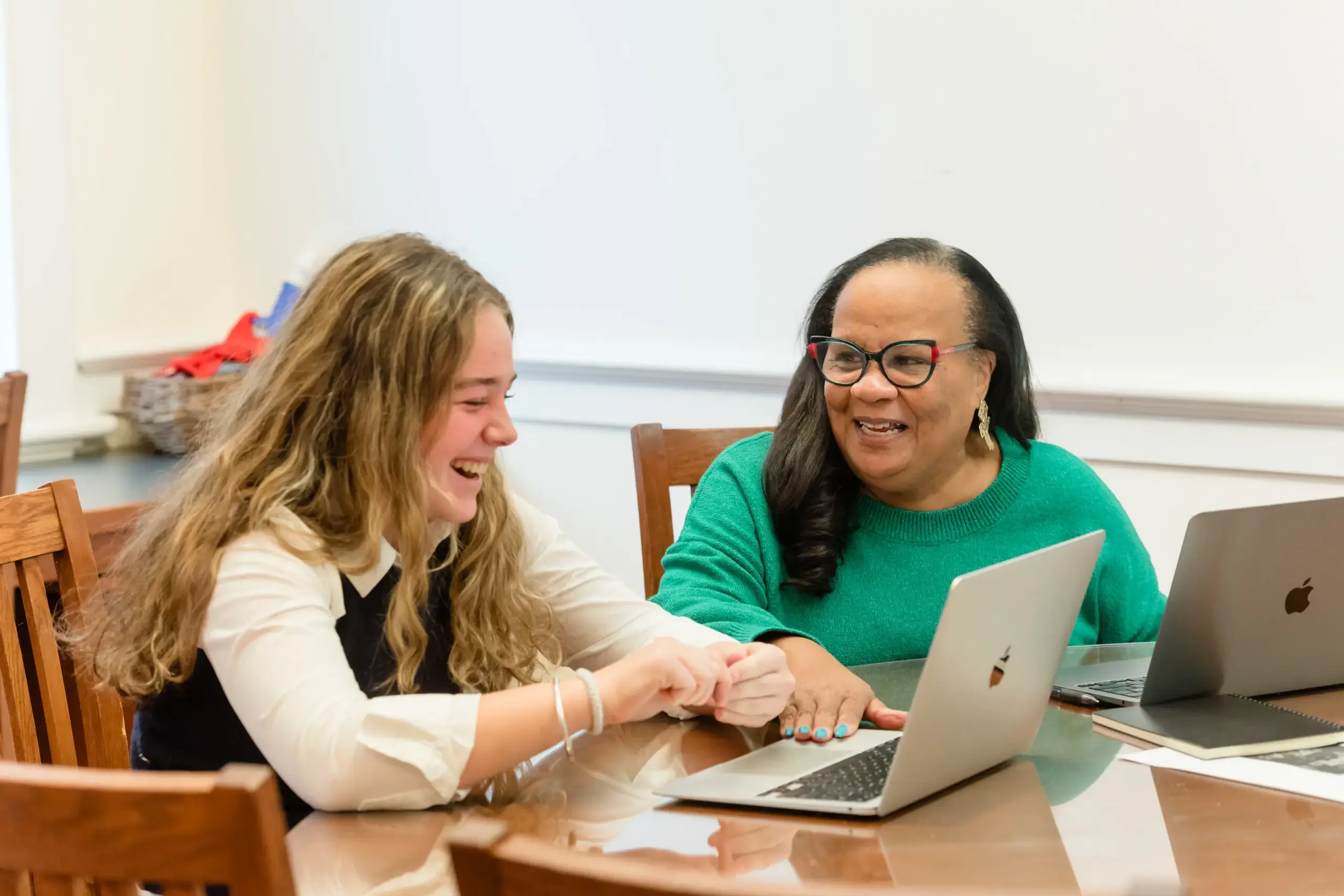 A student and a teacher share a laugh while working on laptops together in a classroom setting. 