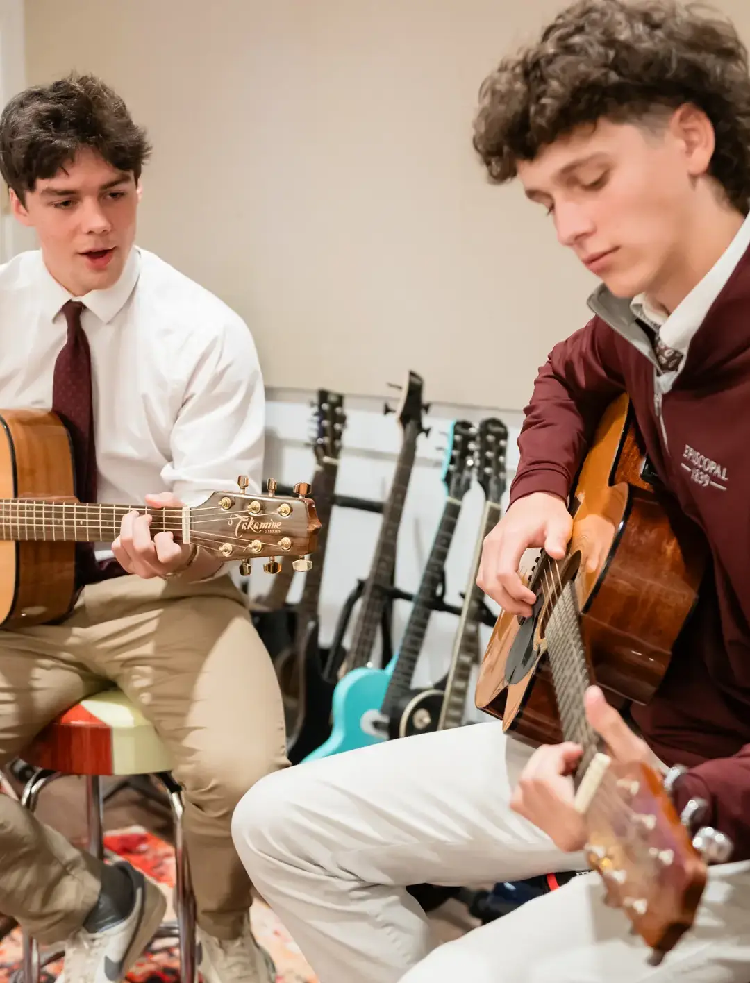 Two students at Episcopal School play acoustic guitars together during a music class, with a rack of additional guitars visible in the background.
