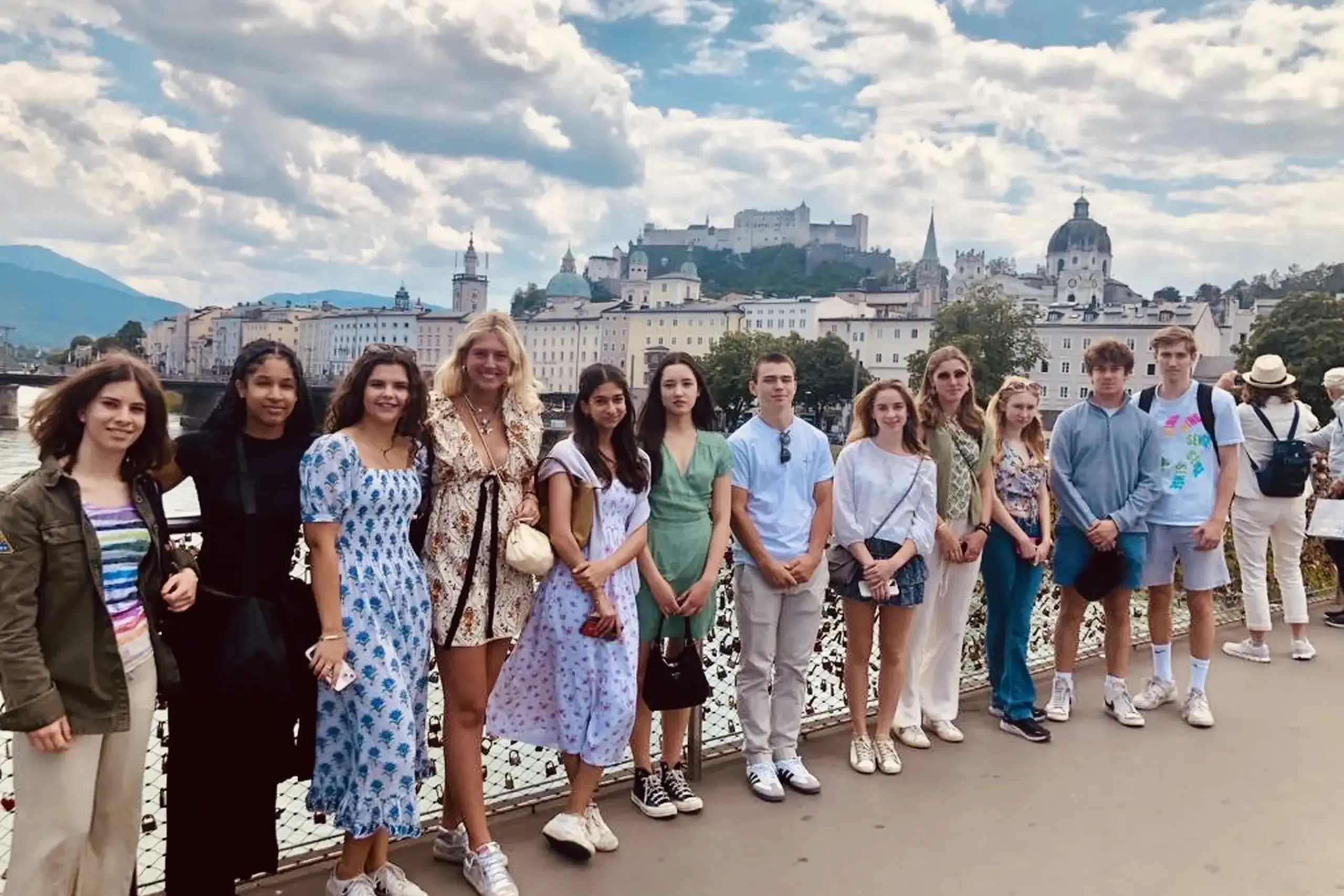 A group of students and one adult stand together on a bridge with a scenic European cityscape in the background, including colorful buildings, church towers, and a historic fortress on a hill.