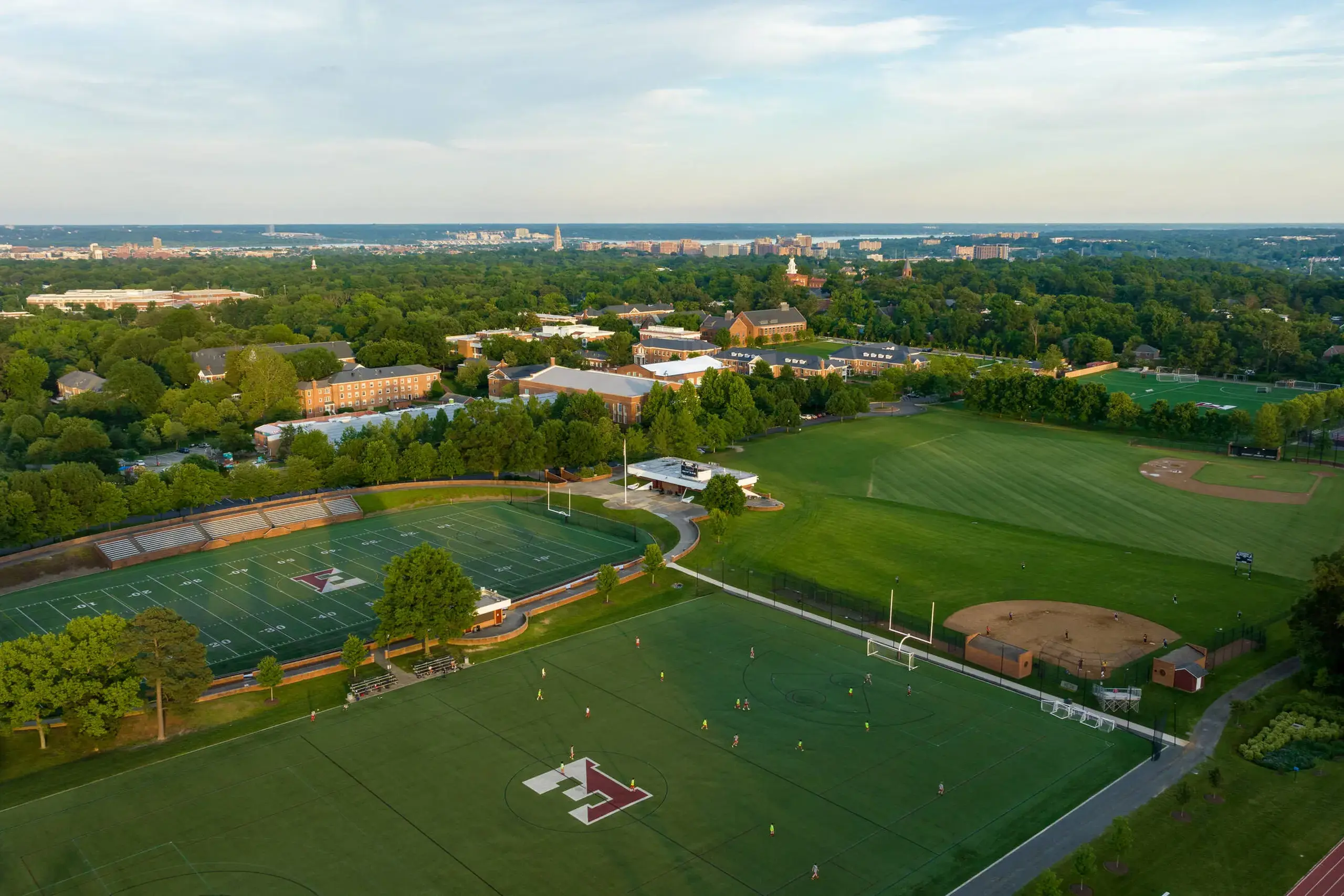 An aerial view of a sprawling campus featuring multiple athletic fields, including a football stadium with bleachers, a soccer field with the school's logo at midfield, and other sporting facilities.