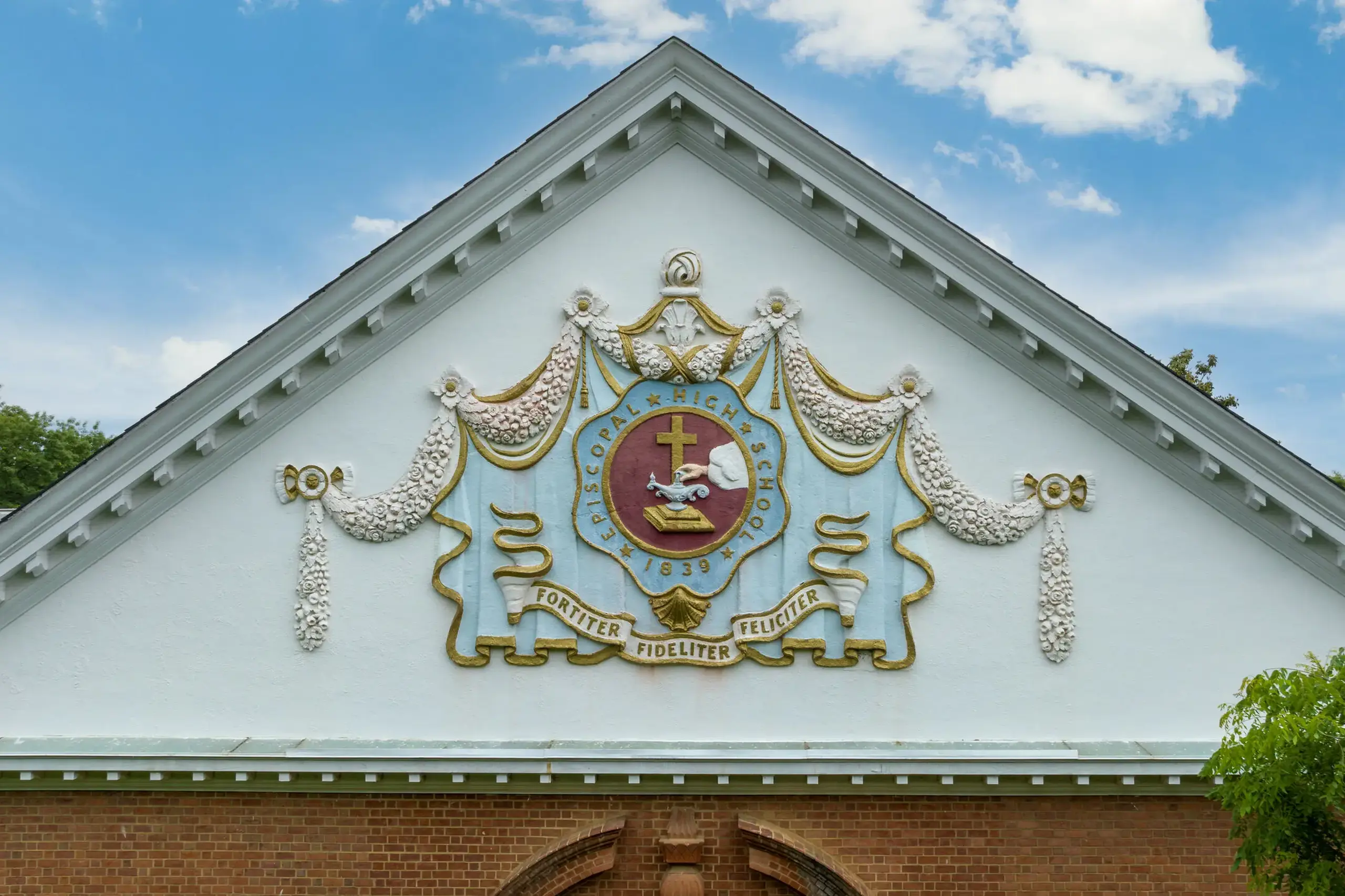 The decorative emblem on the triangular pediment of an Episcopal High School building features an intricate crest with gold and blue detailing. 