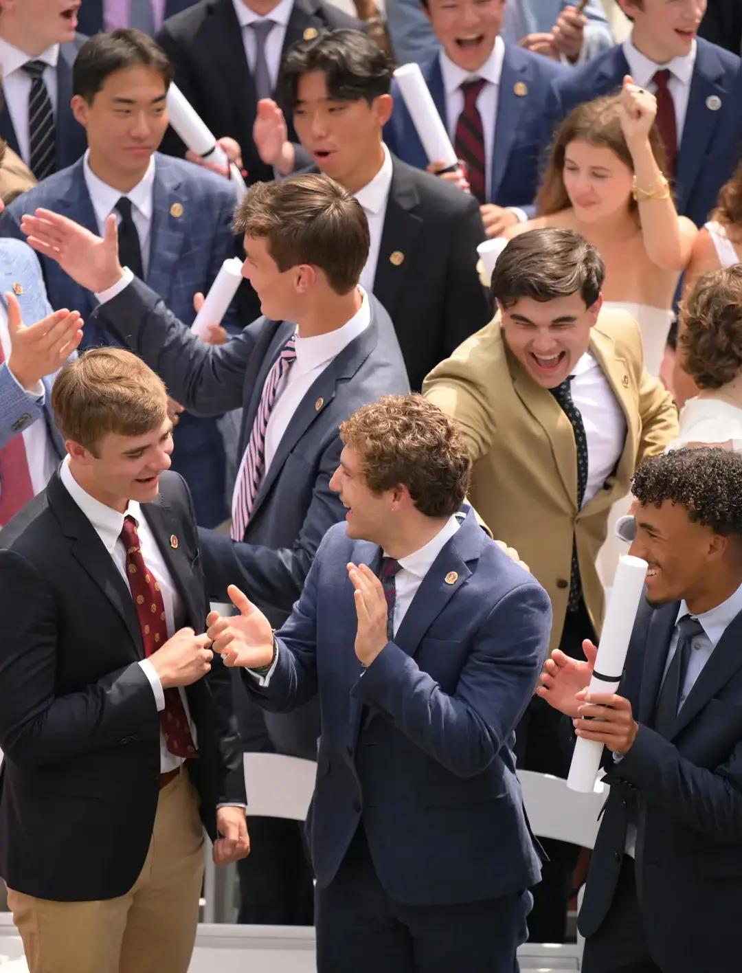 Graduates in formal attire celebrate joyfully, holding diplomas and cheering at an outdoor commencement ceremony.