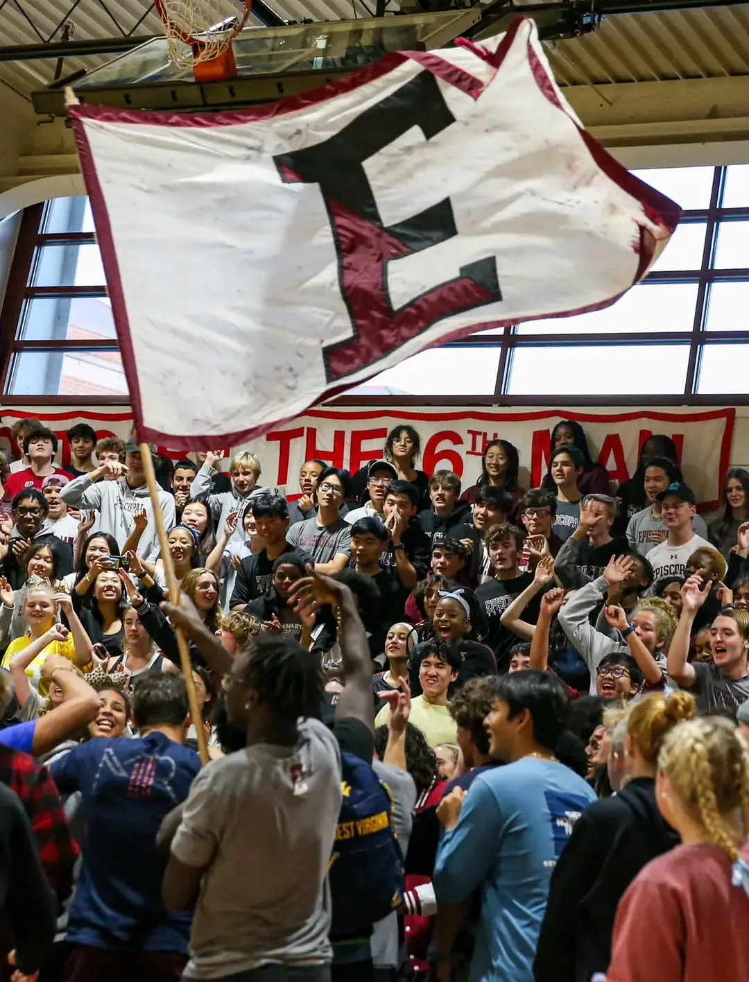 A sea of students fills the gymnasium, cheering, clapping, and waving a large school flag in a display of unstoppable school spirit.