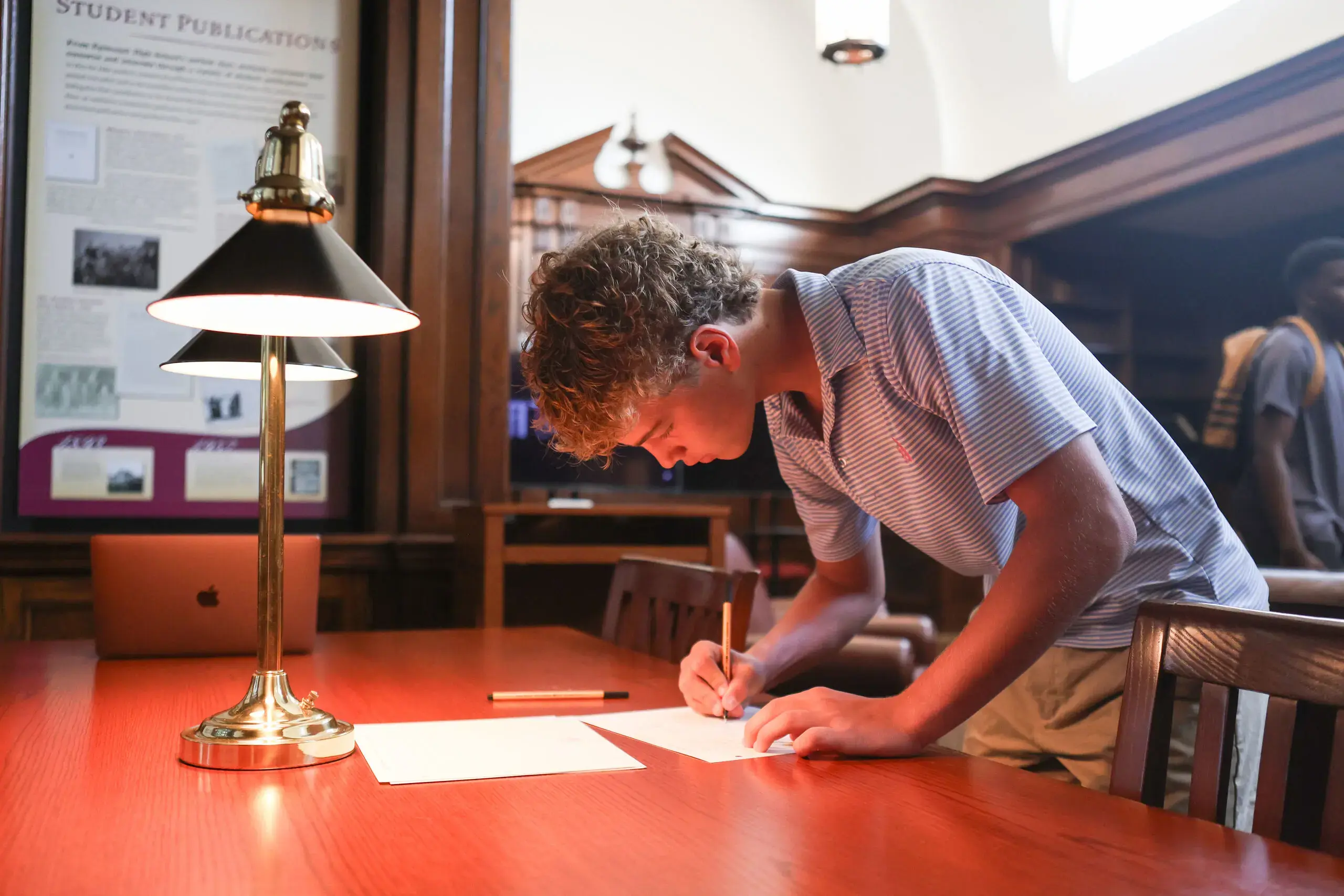 A student with curly blond hair and a striped blue shirt leans over a wooden table, writing on a piece of paper with a focused expression. 