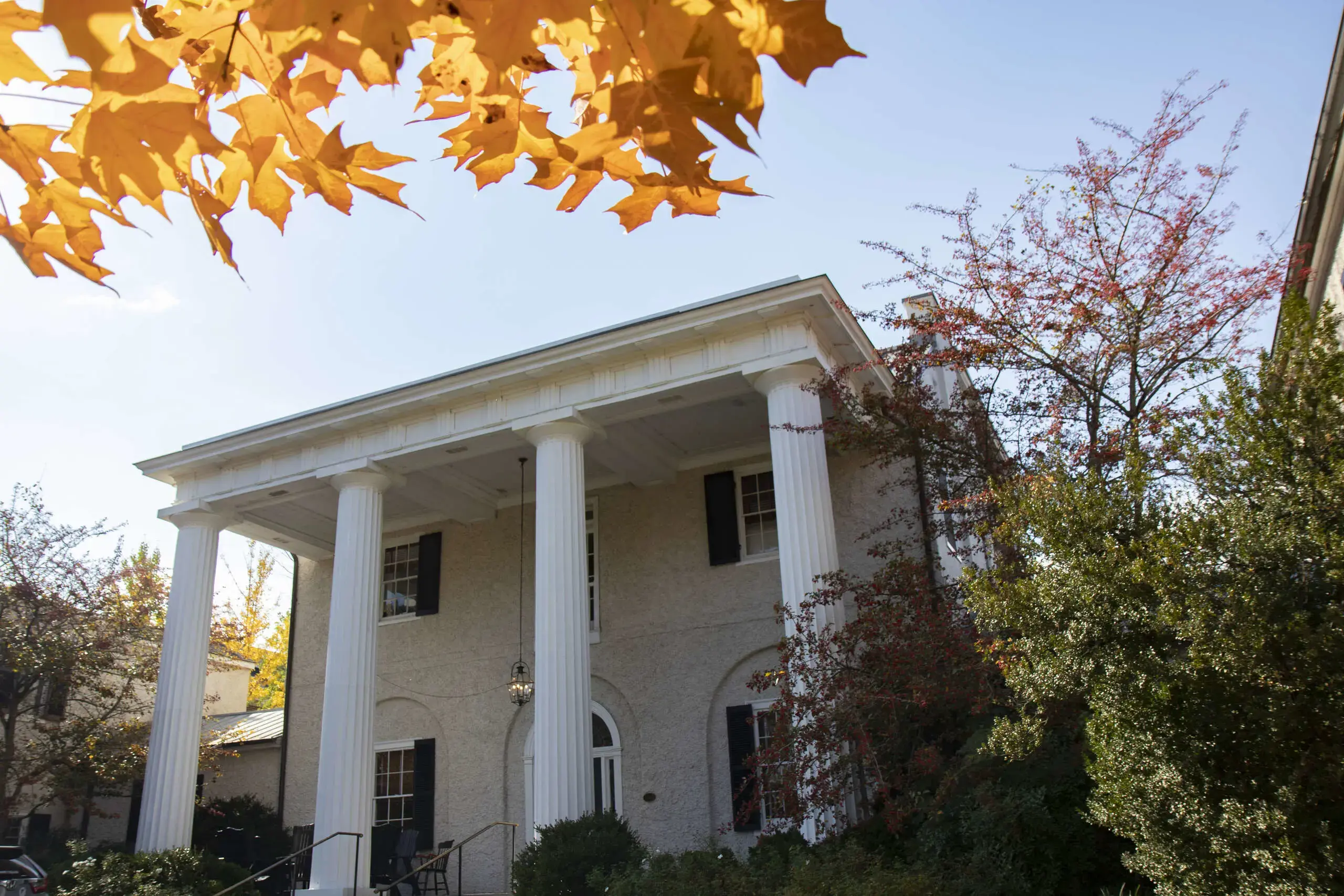 A stately building with tall white columns and classic architecture is framed by vibrant orange autumn leaves in the foreground.