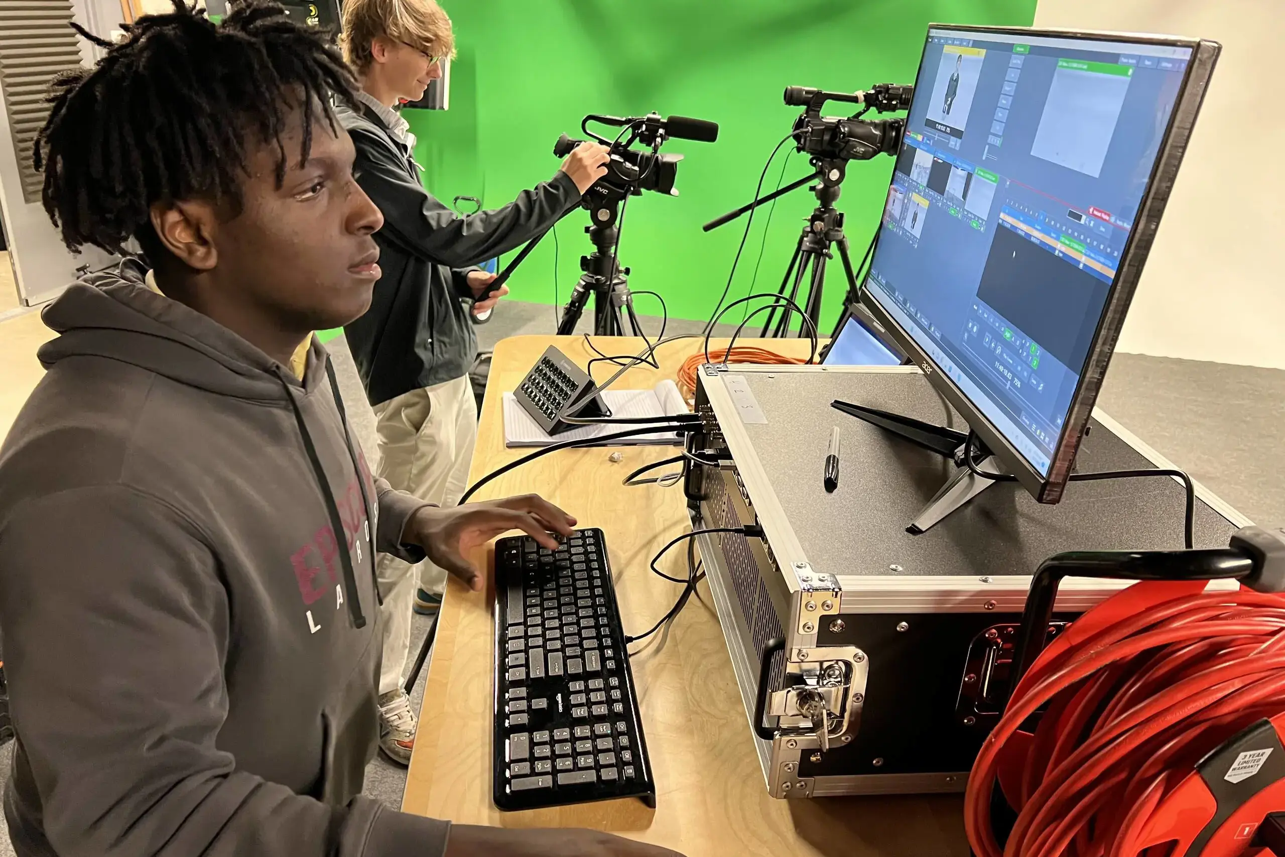 Two students work in a professional video production studio. One student in the foreground operates a computer with video editing software, while the other adjusts a camera on a tripod. 