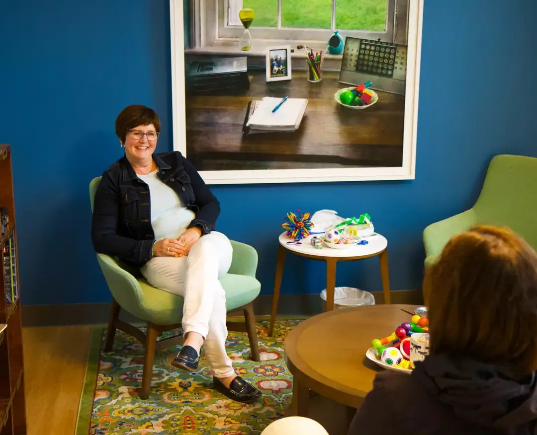 A warm and inviting counseling space where a staff member, seated in a green chair, smiles while engaging in conversation with another individual.