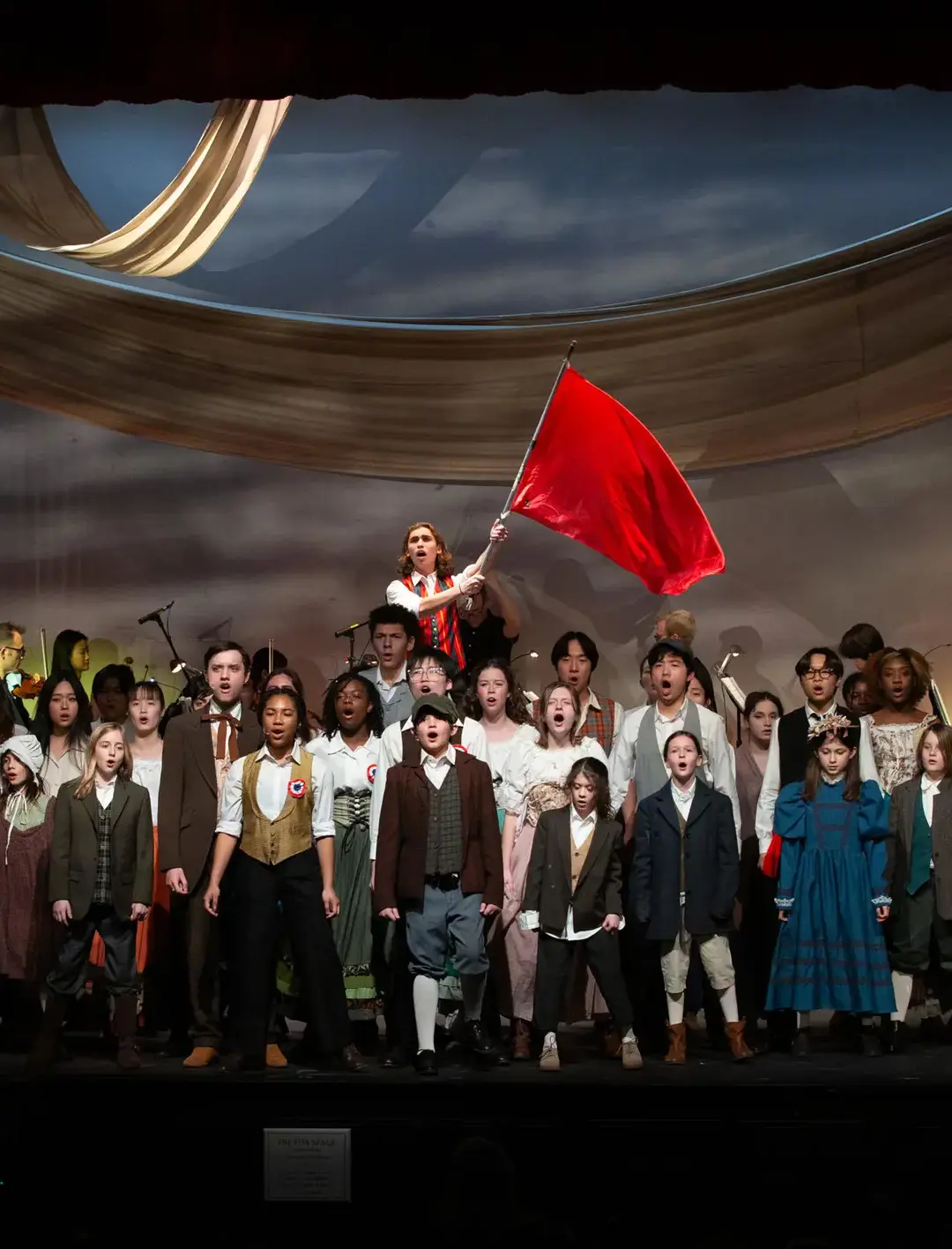 A student theater production performs a dramatic scene on stage, featuring a large ensemble dressed in period costumes.