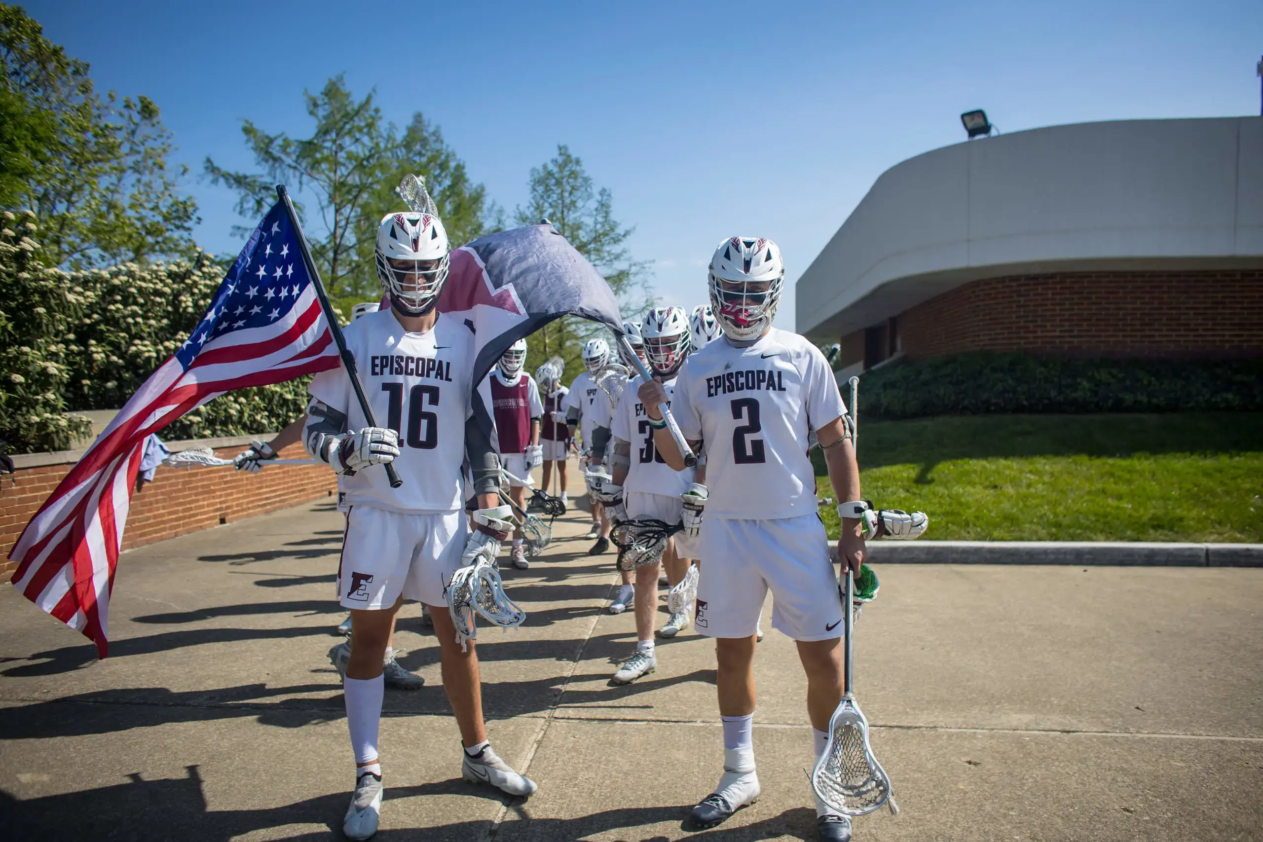 The Episcopal lacrosse team marches onto the field with determination, proudly carrying the American flag and school banner.