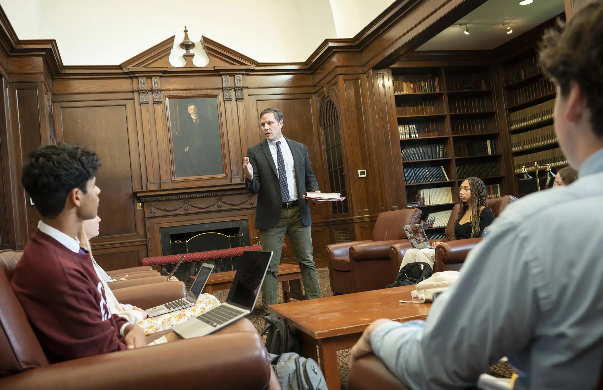 A group of students engaged in a discussion with their teacher in a wood-paneled library, surrounded by bookshelves filled with literature. 
