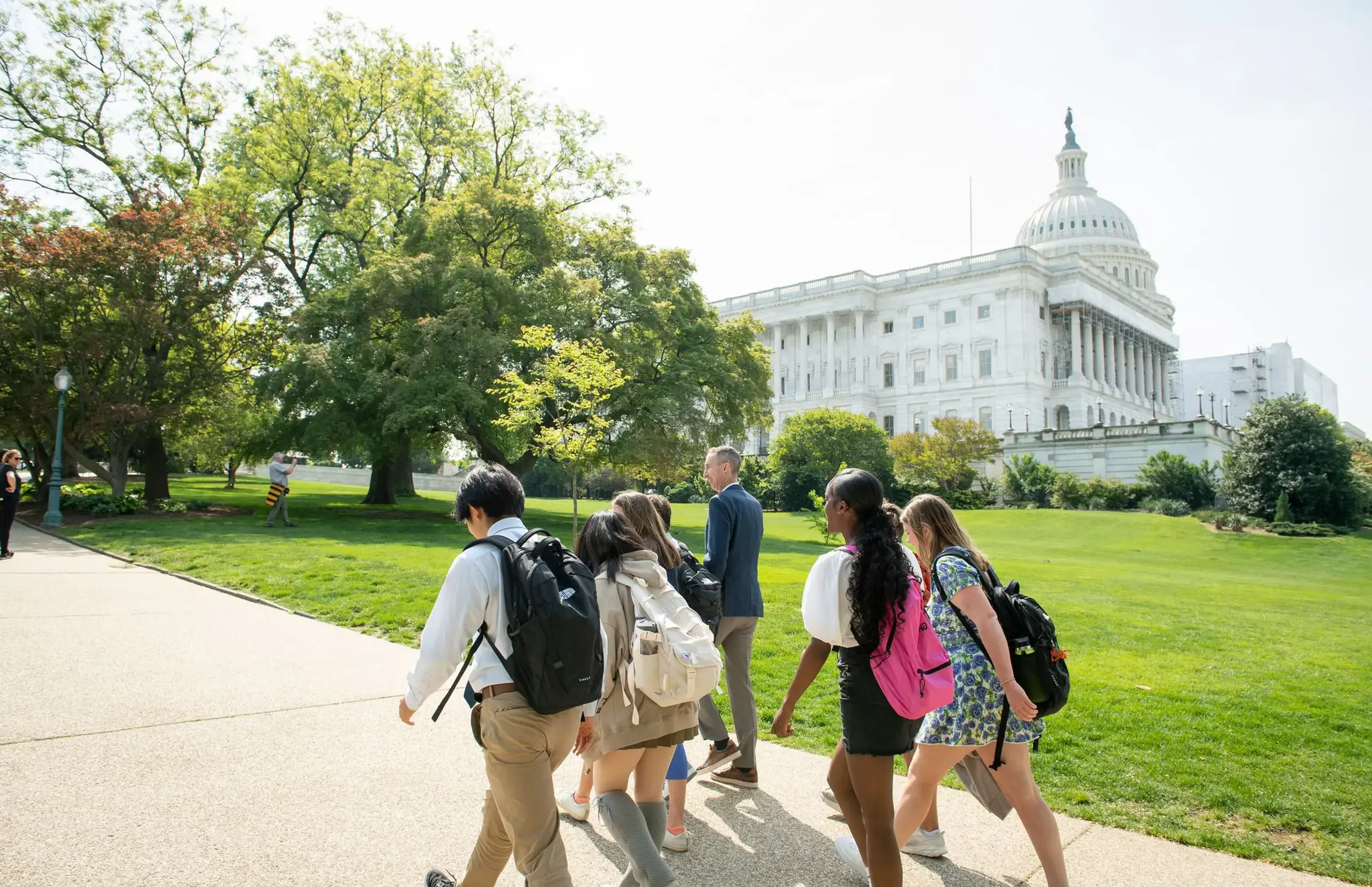 A group of students with backpacks walk along a paved path towards the U.S. Capitol building on a bright, sunny day. 